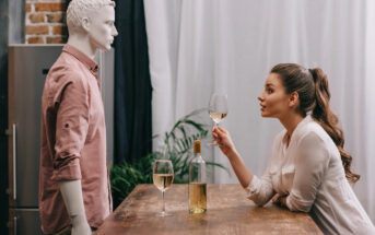 A woman leans on a wooden table holding a glass of white wine, looking intently at a mannequin dressed in a pink shirt. Another glass of wine and a wine bottle sit on the table. A plant and window drapes are in the background.
