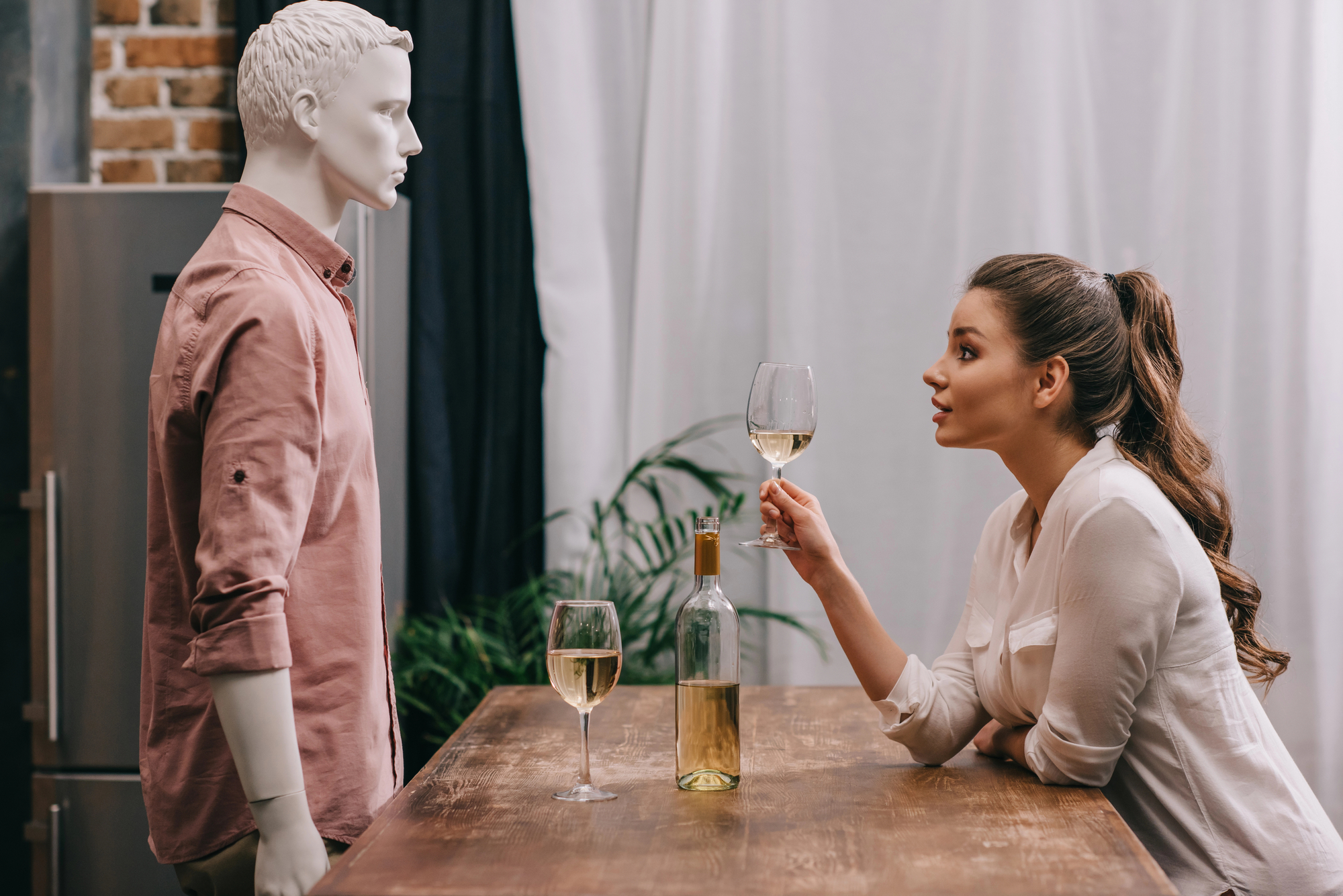 A woman leans on a wooden table holding a glass of white wine, looking intently at a mannequin dressed in a pink shirt. Another glass of wine and a wine bottle sit on the table. A plant and window drapes are in the background.