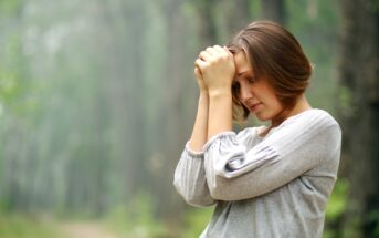 A woman with short brown hair stands in a forest, wearing a light gray sweater. She has a thoughtful or solemn expression, resting her forehead on her clasped hands. The background is filled with tall trees and a narrow path, blurred to create a peaceful ambiance.