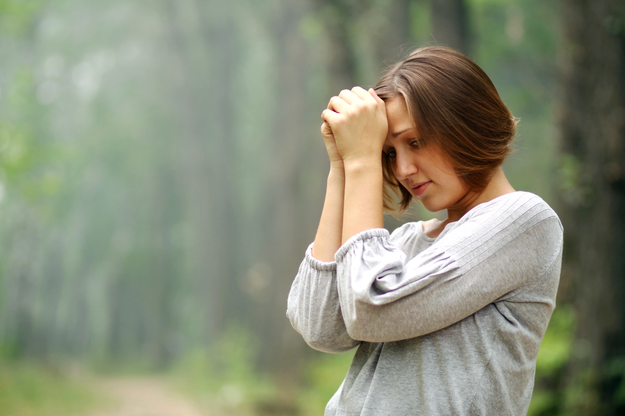 A woman with short brown hair stands in a forest, wearing a light gray sweater. She has a thoughtful or solemn expression, resting her forehead on her clasped hands. The background is filled with tall trees and a narrow path, blurred to create a peaceful ambiance.