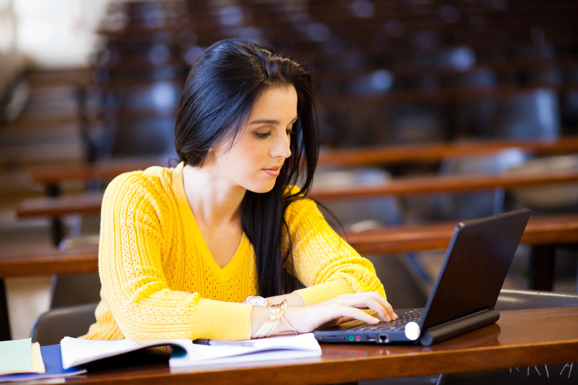 A woman with long dark hair and a yellow sweater sits at a desk typing on a laptop in a classroom. Open notebooks and papers are on the desk in front of her. Rows of empty chairs and desks are visible in the background.