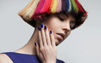 A person with a distinctive short bob haircut dyed in a vibrant rainbow of colors touches their face with a hand that has cobalt blue nail polish. They are wearing a matching blue top and looking down, creating a contemplative pose against a plain background.