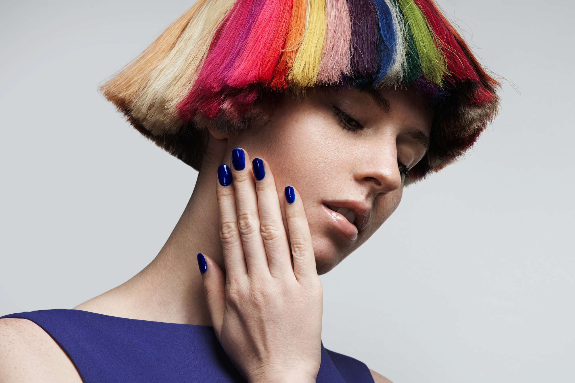 A person with a distinctive short bob haircut dyed in a vibrant rainbow of colors touches their face with a hand that has cobalt blue nail polish. They are wearing a matching blue top and looking down, creating a contemplative pose against a plain background.