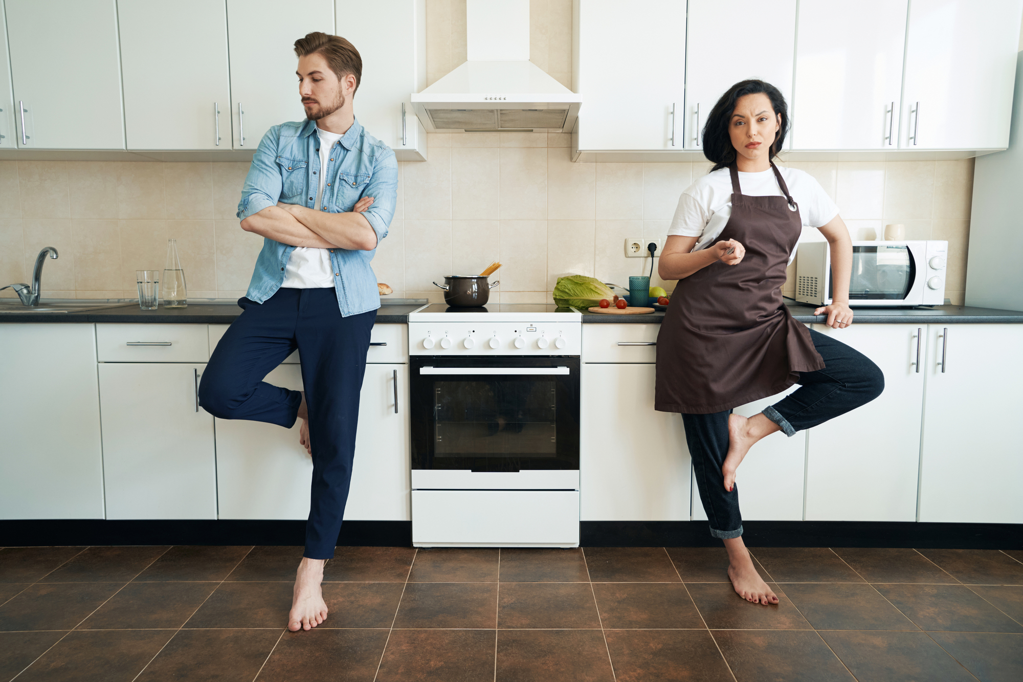 A man and a woman are in a kitchen, both standing barefoot. The man, wearing a denim shirt over a white t-shirt and dark pants, has his arms crossed and leans against the counter. The woman, in a brown apron and jeans, is holding a spoon and standing in a yoga pose.