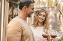 A man and a woman enjoying glasses of red wine outdoors. They are standing close to each other, smiling and making eye contact. The setting appears to be a sunny day with trees and a building in the background. Both are dressed casually.