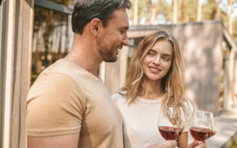 A man and a woman enjoying glasses of red wine outdoors. They are standing close to each other, smiling and making eye contact. The setting appears to be a sunny day with trees and a building in the background. Both are dressed casually.