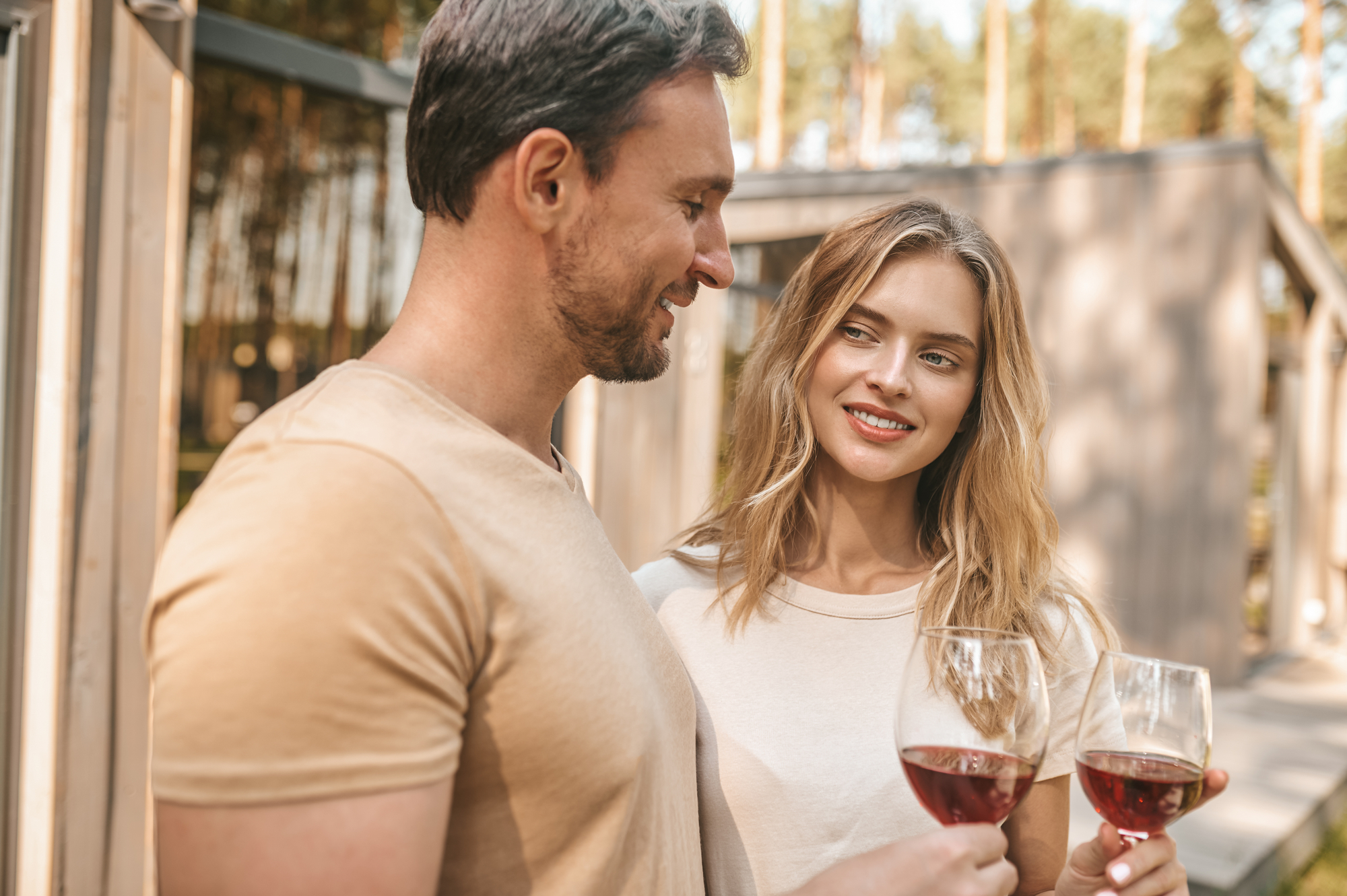 A man and a woman enjoying glasses of red wine outdoors. They are standing close to each other, smiling and making eye contact. The setting appears to be a sunny day with trees and a building in the background. Both are dressed casually.