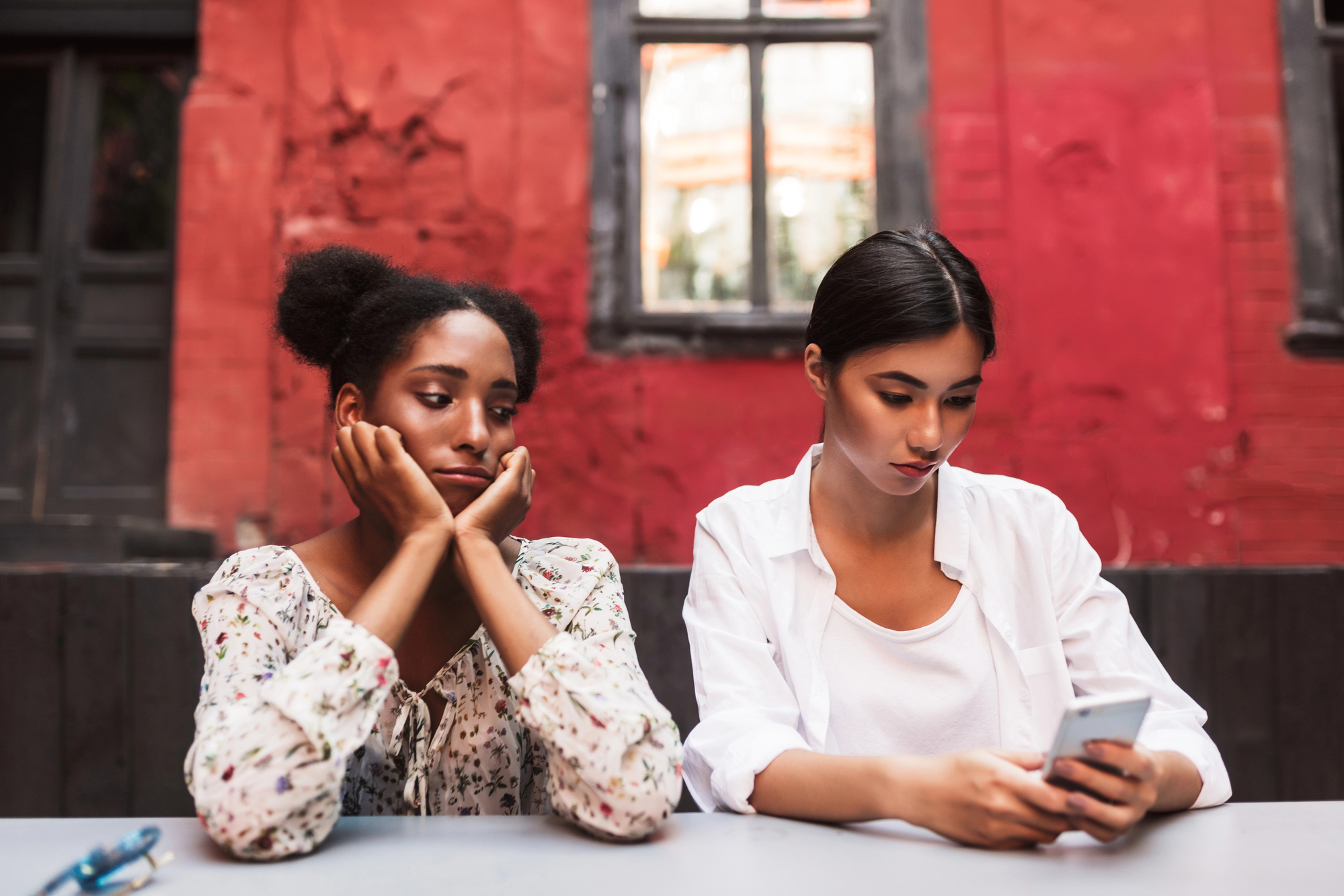 Two women sitting at a table in an outdoor cafe. One woman, with her hands on her face, looks bored or annoyed while the other, in a white shirt, is focused on her smartphone. They are in front of a red wall with a window and a closed door.