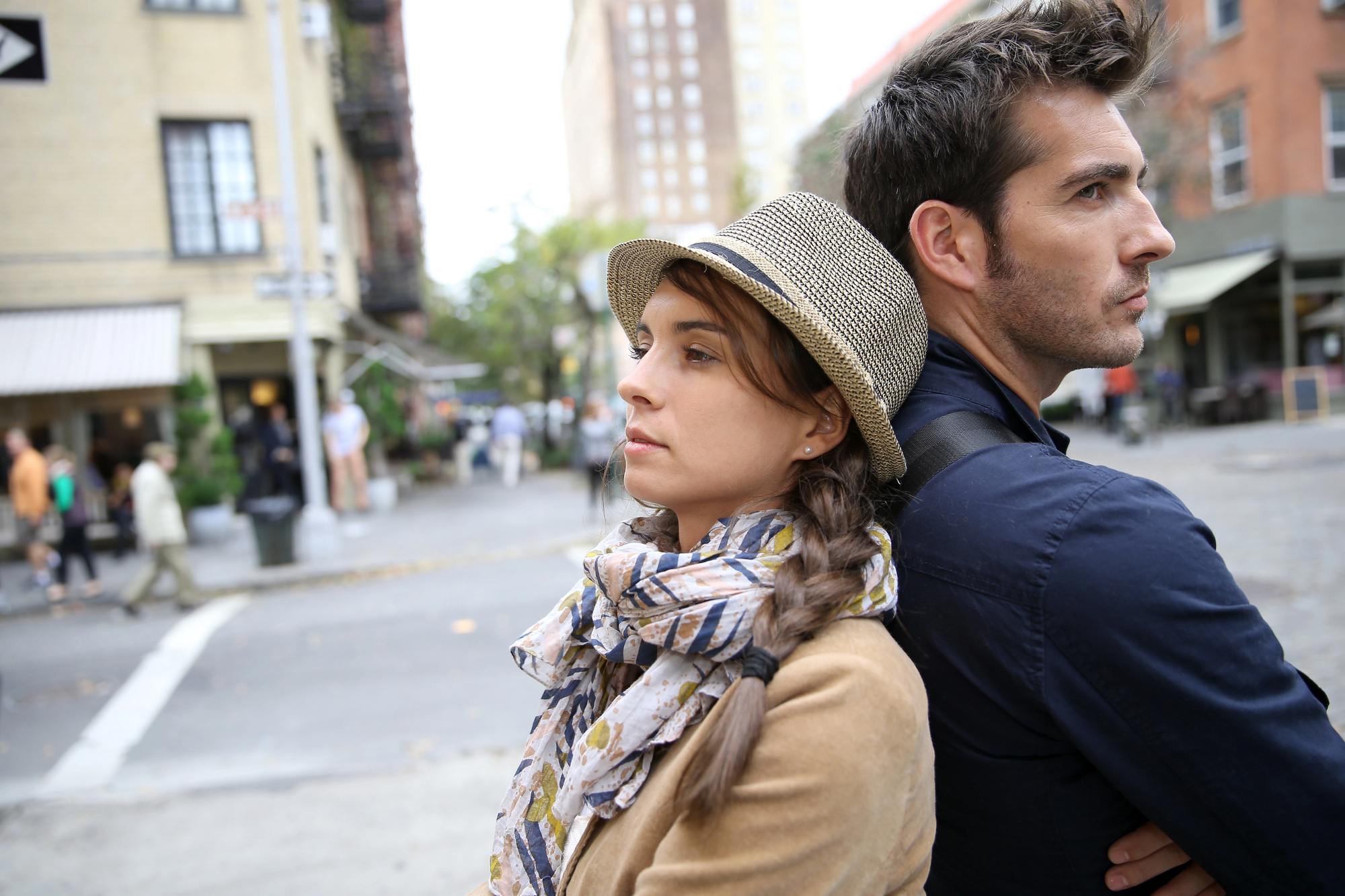 A woman wearing a hat and scarf stands back-to-back with a man in a blue shirt on a city street. Both look distant and thoughtful. The background includes buildings, trees, and pedestrians.