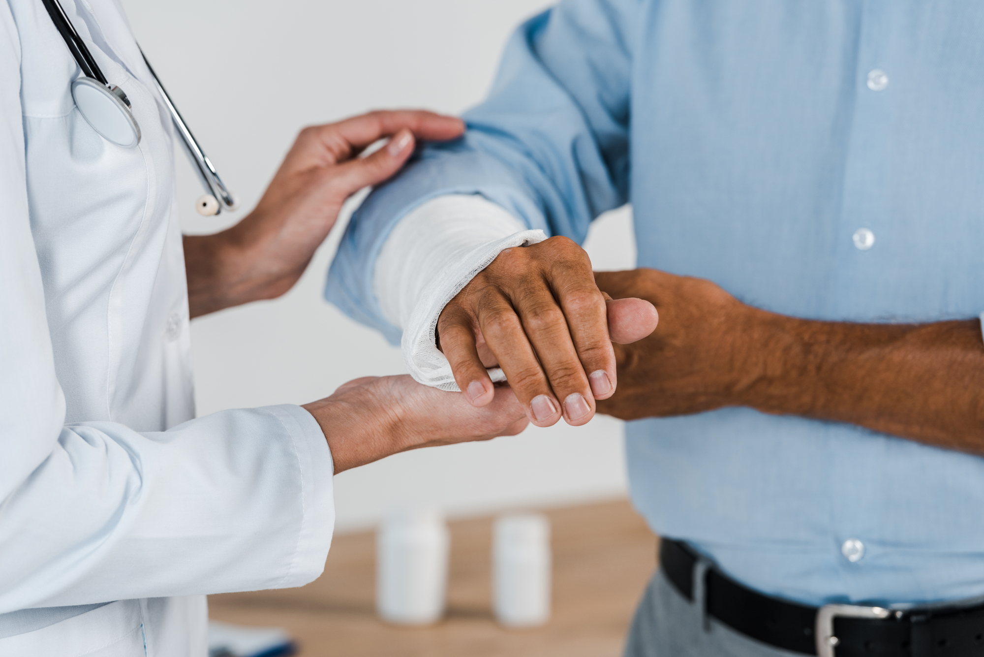 A doctor in a white coat is examining a patient's arm, which is wrapped in a bandage. The patient is wearing a light blue shirt, and their hands are gently held by the doctor. The background is a plain, softly lit room.