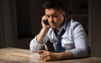 A man sitting at a wooden table, wearing a light blue shirt and a loosened dark blue tie, holds a smartphone to his ear with a serious expression. His other hand is resting on the table, holding a glass of brown liquid, possibly whiskey. The background is dimly lit.