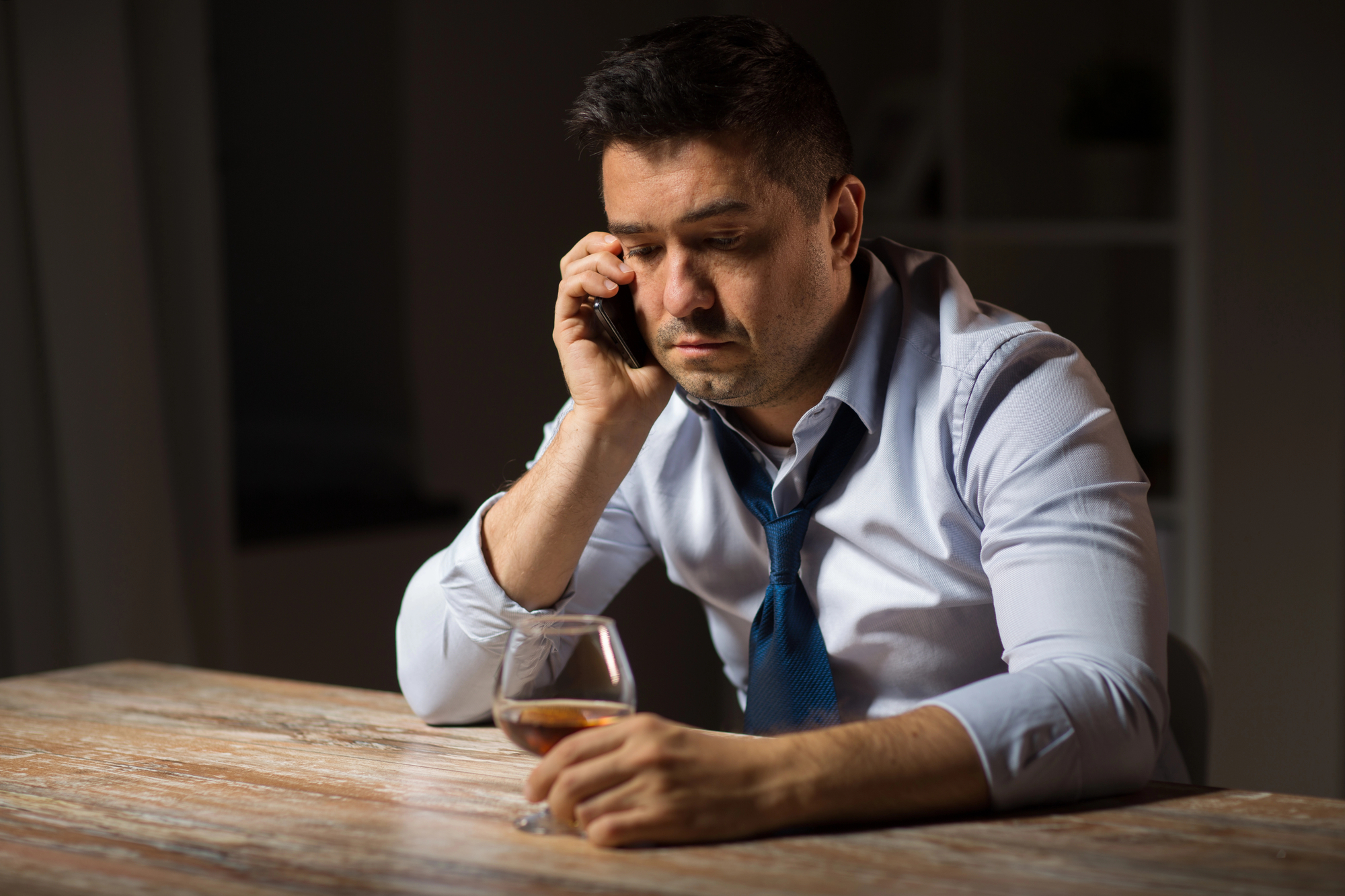 A man sitting at a wooden table, wearing a light blue shirt and a loosened dark blue tie, holds a smartphone to his ear with a serious expression. His other hand is resting on the table, holding a glass of brown liquid, possibly whiskey. The background is dimly lit.