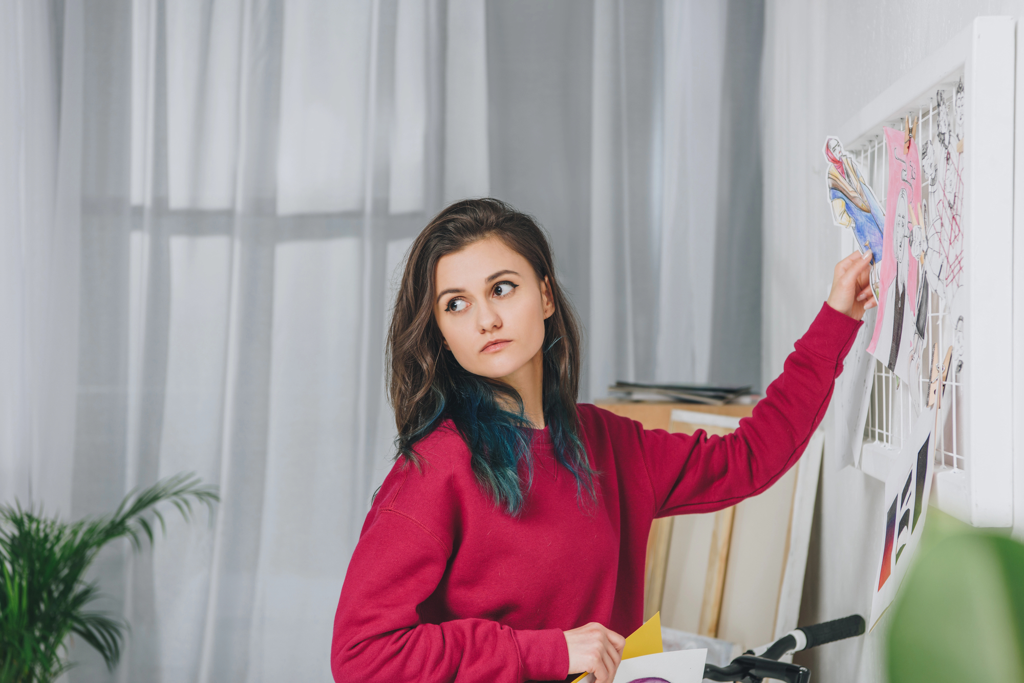 A young woman with long brown hair, wearing a red sweater, stands next to a white pinboard covered with various pictures and notes. She is looking to her right with a thoughtful expression. A large window with sheer curtains and a potted plant are in the background.