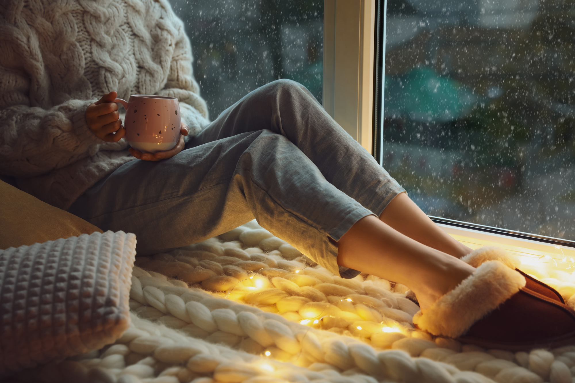 A person wearing a cozy sweater and jeans sits by a window holding a mug. Outside, it appears to be raining. The person's feet, in slippers, rest on a knitted blanket decorated with string lights, creating a warm and relaxing atmosphere.