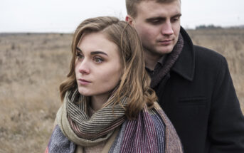 A woman and man stand close together in a barren, brown field. The woman, with light brown hair and a striped scarf, gazes forward with a serious expression. The man, wearing a dark coat, stands slightly behind her, looking down. Both appear contemplative.