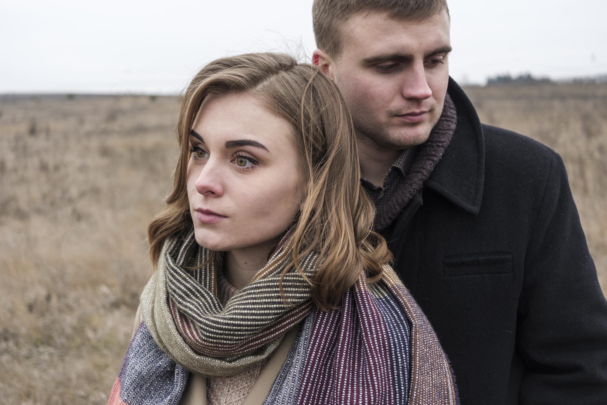 A woman and man stand close together in a barren, brown field. The woman, with light brown hair and a striped scarf, gazes forward with a serious expression. The man, wearing a dark coat, stands slightly behind her, looking down. Both appear contemplative.