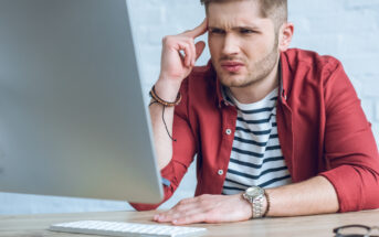 A young man with short hair and a beard, wearing a red shirt over a striped t-shirt, sits at a desk, looking at a computer screen with a confused expression. His left hand is touching his temple, and a keyboard and glasses case are on the desk in front of him.