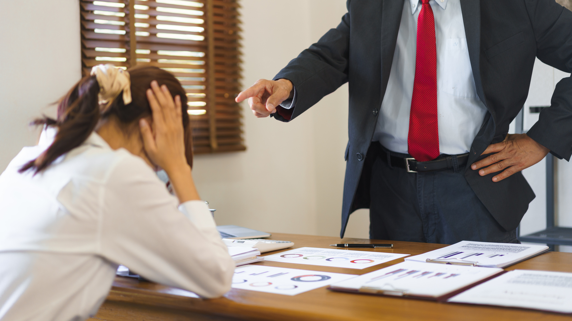 A man in a suit and red tie is standing and pointing at a woman sitting at a desk with her head in her hands, appearing stressed. The desk has several charts and documents spread out. Wooden blinds and a beige wall are in the background.