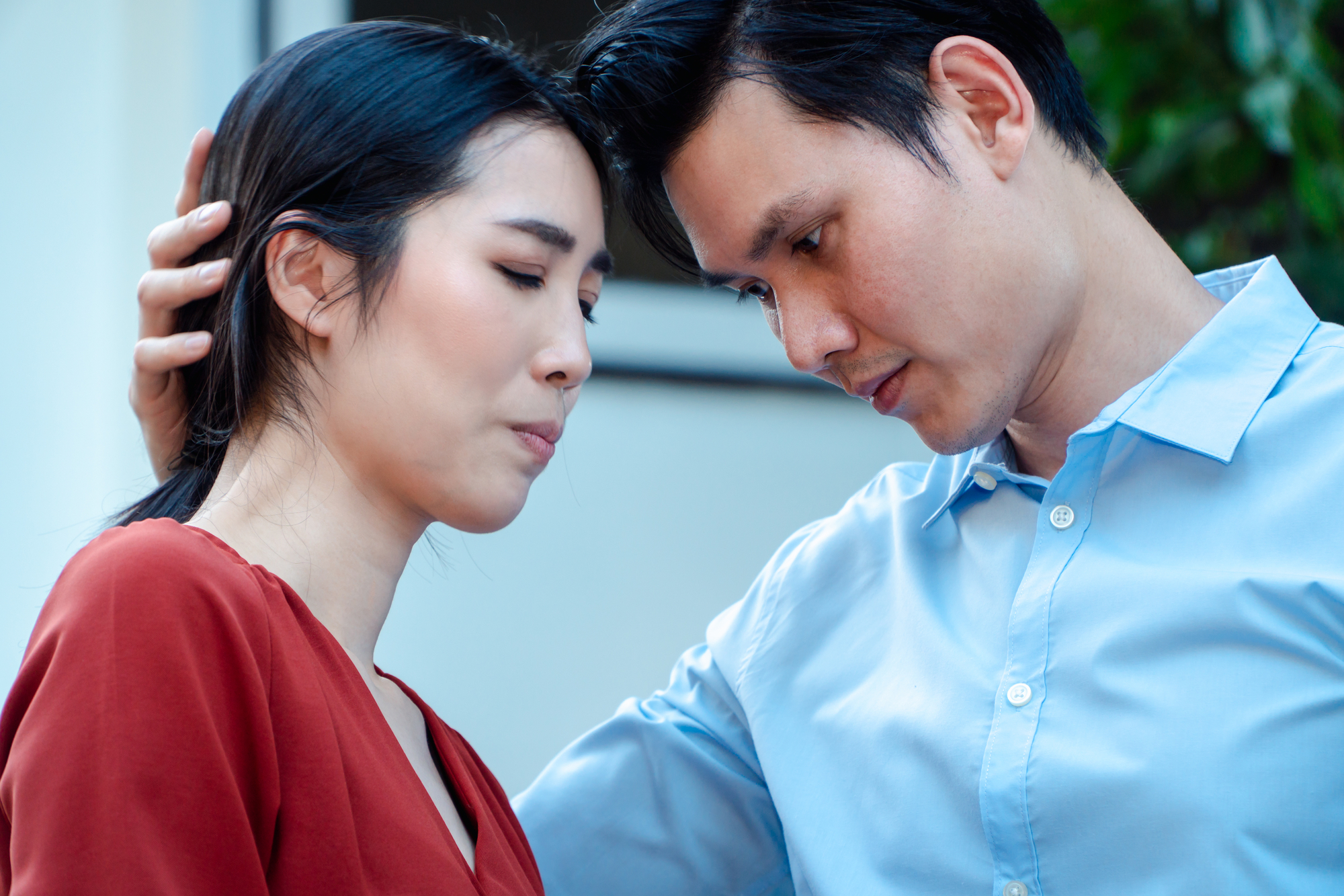 A man wearing a light blue shirt gently cradles the head of a woman in a red blouse. Both appear to be in a moment of deep emotion. The man rests his forehead against the woman's as she looks down. The background is blurred, suggesting an outdoor setting.