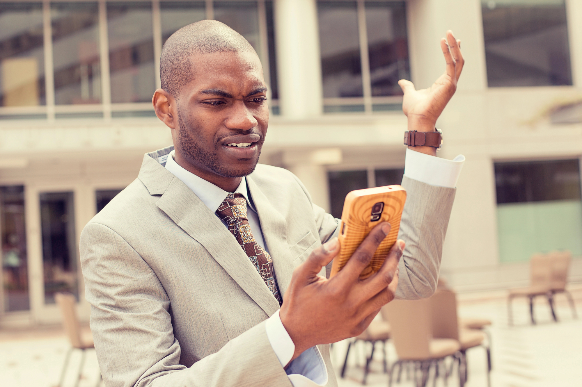 A man in a gray suit stands outdoors in front of a modern building, holding a smartphone in his left hand. He appears puzzled or frustrated, with his right hand raised in a questioning gesture and a furrowed brow.