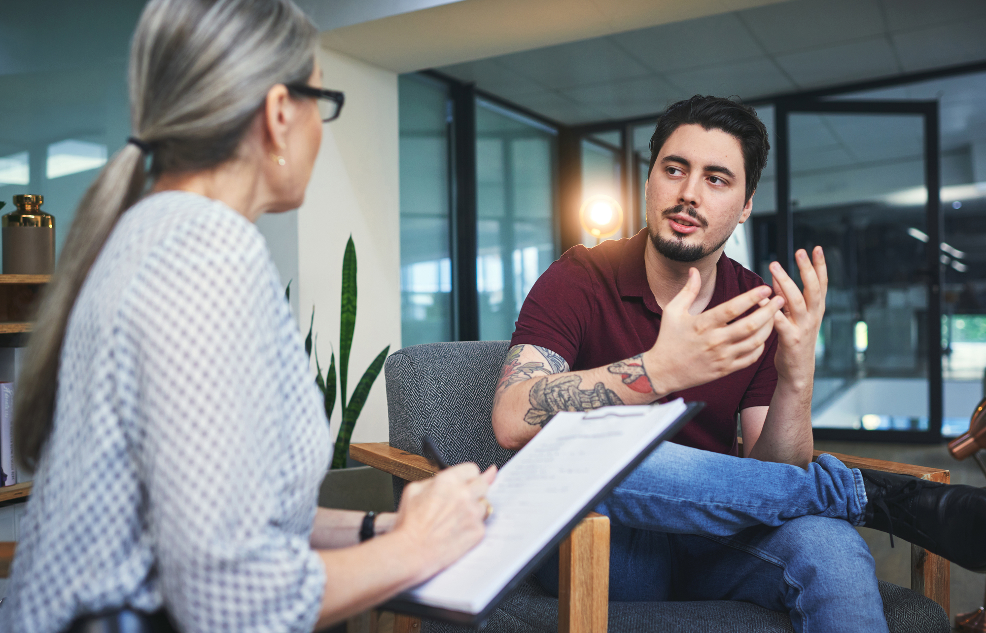 A man with tattoos on his arms is sitting on a chair gesturing with his hands while talking to a woman with gray hair in a ponytail who is holding a clipboard. They appear to be in a modern office or counseling setting with large windows in the background.