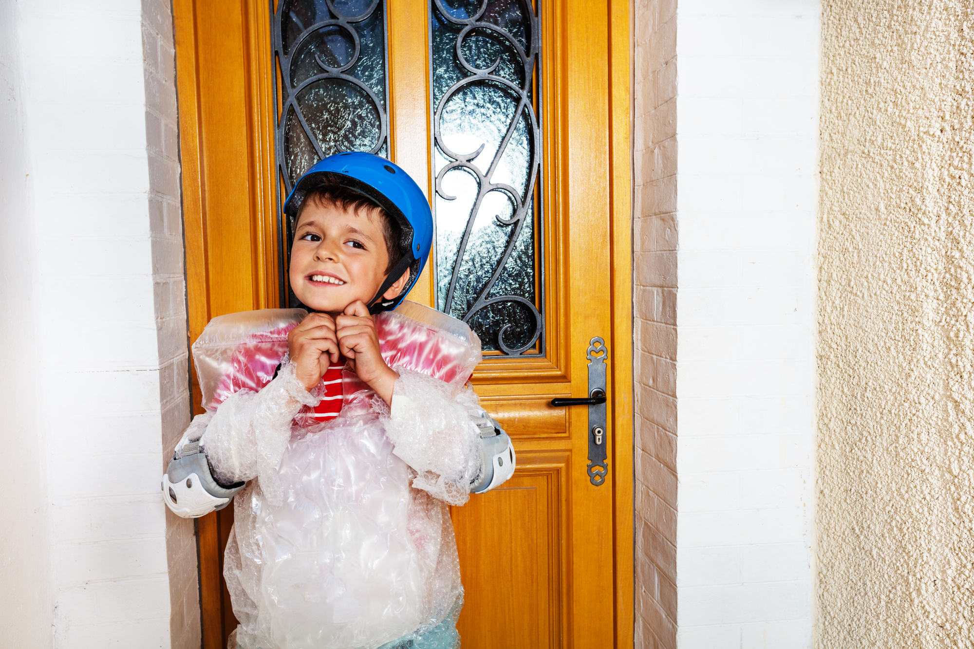 A smiling child stands in front of a wooden door, wearing a blue helmet and protective gear made of bubble wrap. The child appears excited and ready for an adventure, with arm pads visible on both arms. The door behind them has decorative metalwork on the glass panels.