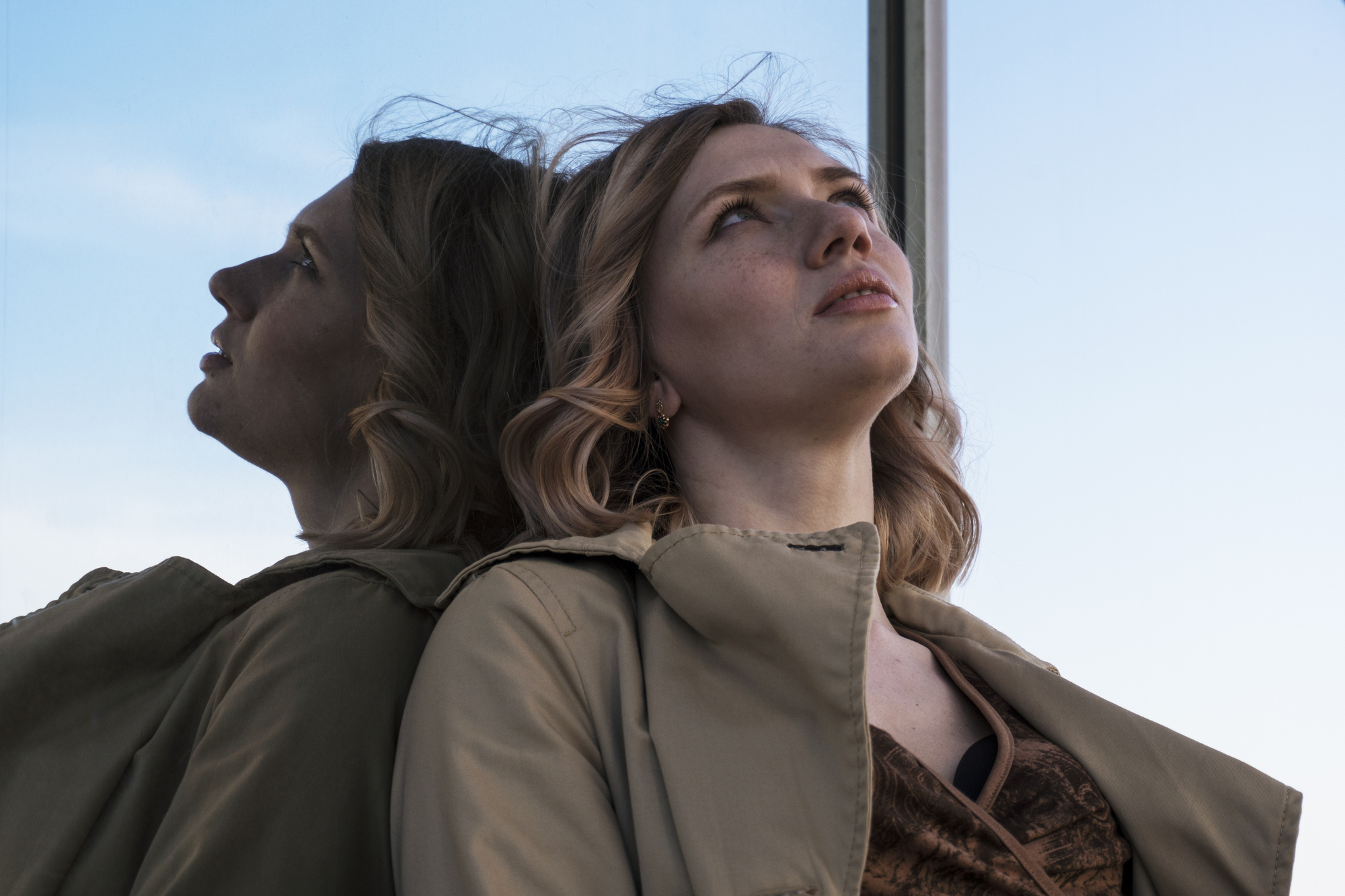 A woman with blonde hair gazes upward while leaning against a reflective glass surface. Her reflection is visible behind her. She is wearing a beige coat and a brown top. The sky is clear and blue in the background.