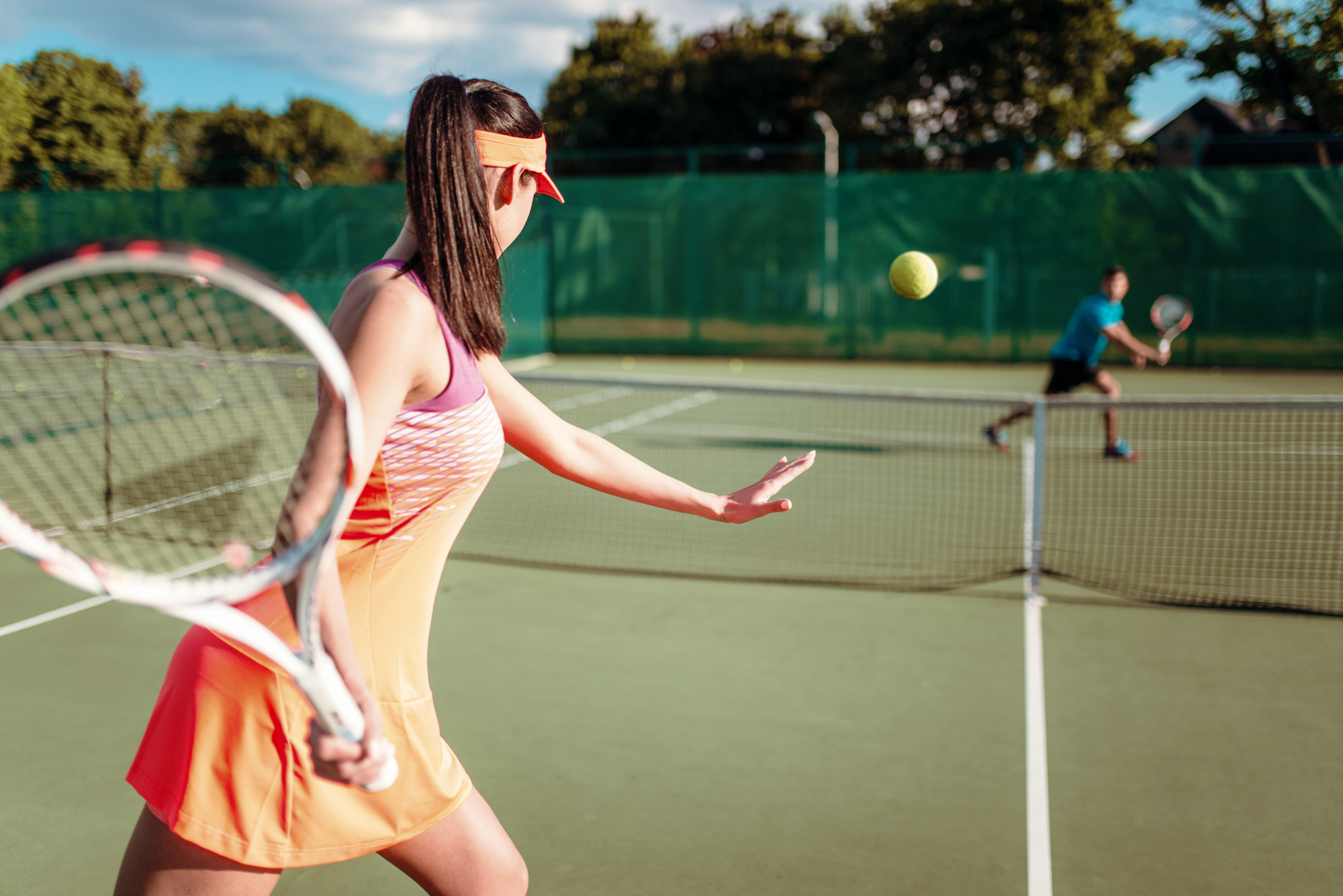 A woman in an orange tennis outfit and visor is preparing to return a tennis ball with her racquet. She is on a tennis court, with a male player in the background about to hit the ball. The background includes trees and a green fence.