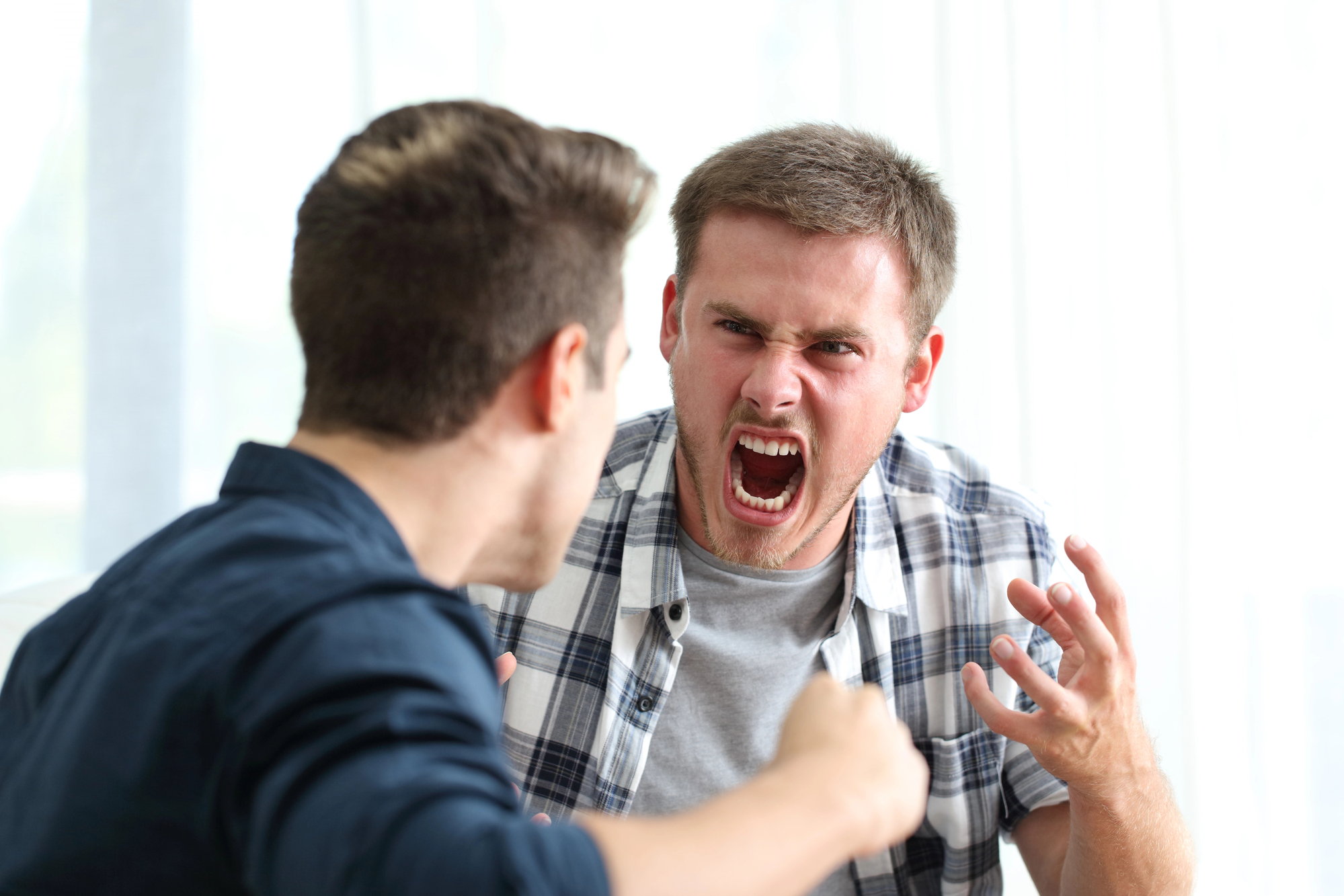 Two men are in a heated argument indoors. The man on the right is yelling with his mouth wide open, glaring angrily, and raising his hands. The man on the left, seen from behind, appears to be responding intensely. Both men have short, brown hair.