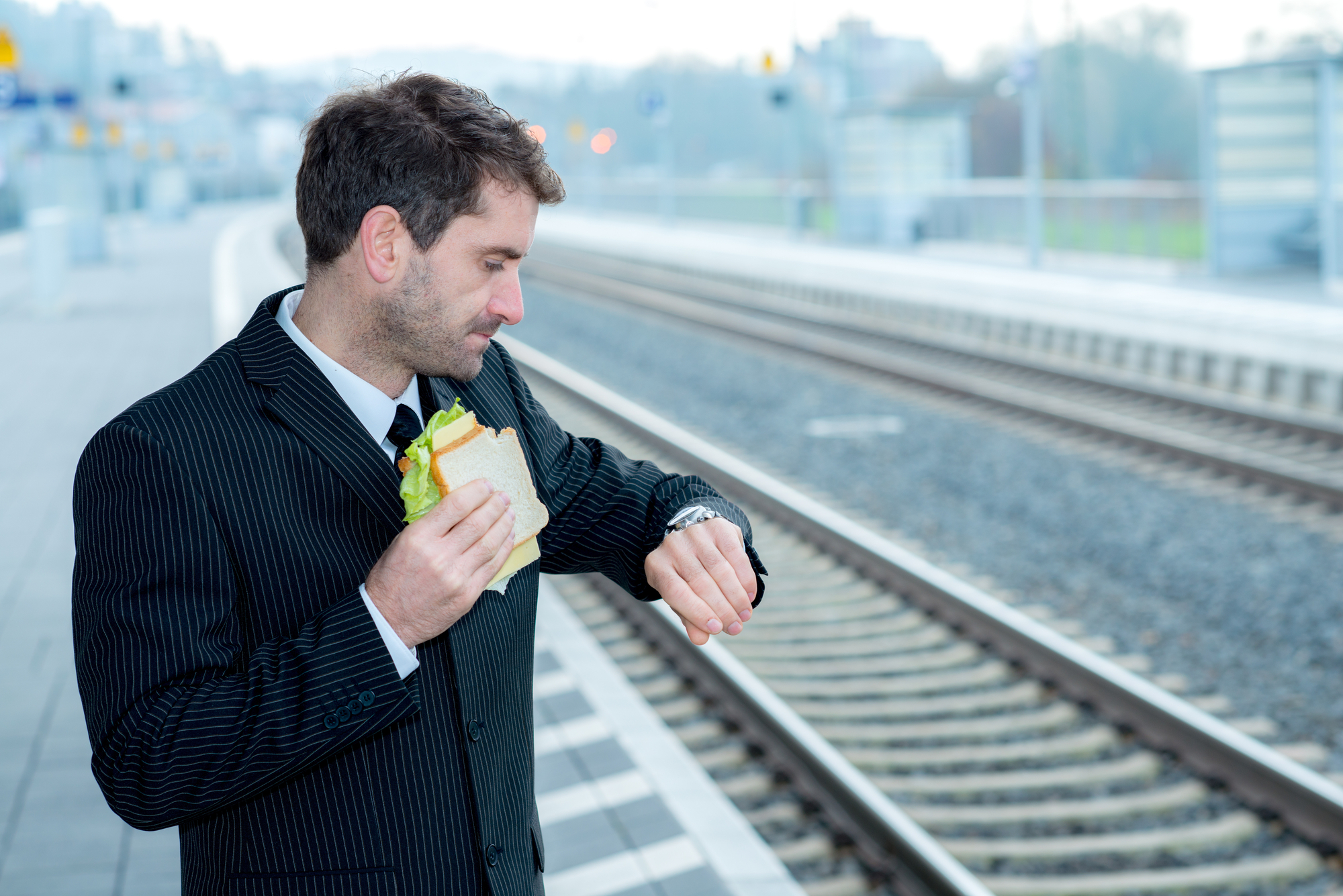 A man in a suit stands on a train platform, holding a sandwich in one hand while looking at his wristwatch with a concerned expression. The train tracks and platform are visible in the background under a cloudy sky.