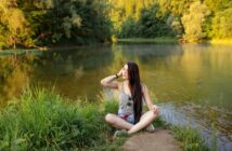 A woman with long dark hair sits cross-legged on a patch of grass near a calm river, surrounded by lush, green trees. She is wearing a grey tank top and shorts, looking up while touching her hair and sunglasses rest on her chest. The scene is peaceful and sunlit.