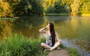 A woman with long dark hair sits cross-legged on a patch of grass near a calm river, surrounded by lush, green trees. She is wearing a grey tank top and shorts, looking up while touching her hair and sunglasses rest on her chest. The scene is peaceful and sunlit.