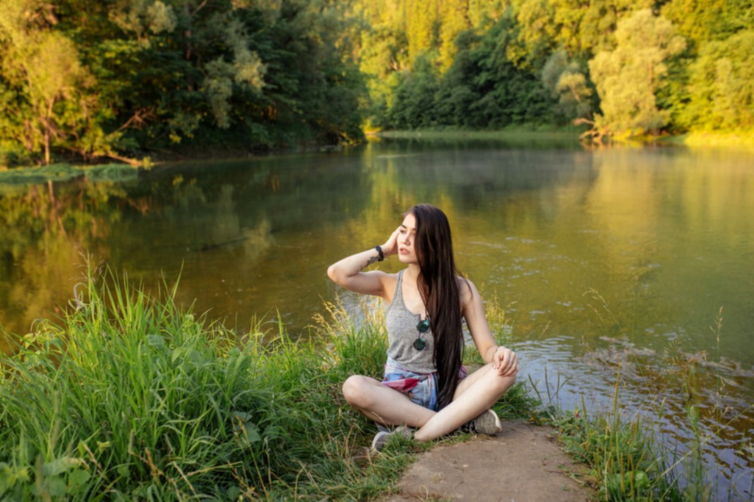 A woman with long dark hair sits cross-legged on a patch of grass near a calm river, surrounded by lush, green trees. She is wearing a grey tank top and shorts, looking up while touching her hair and sunglasses rest on her chest. The scene is peaceful and sunlit.