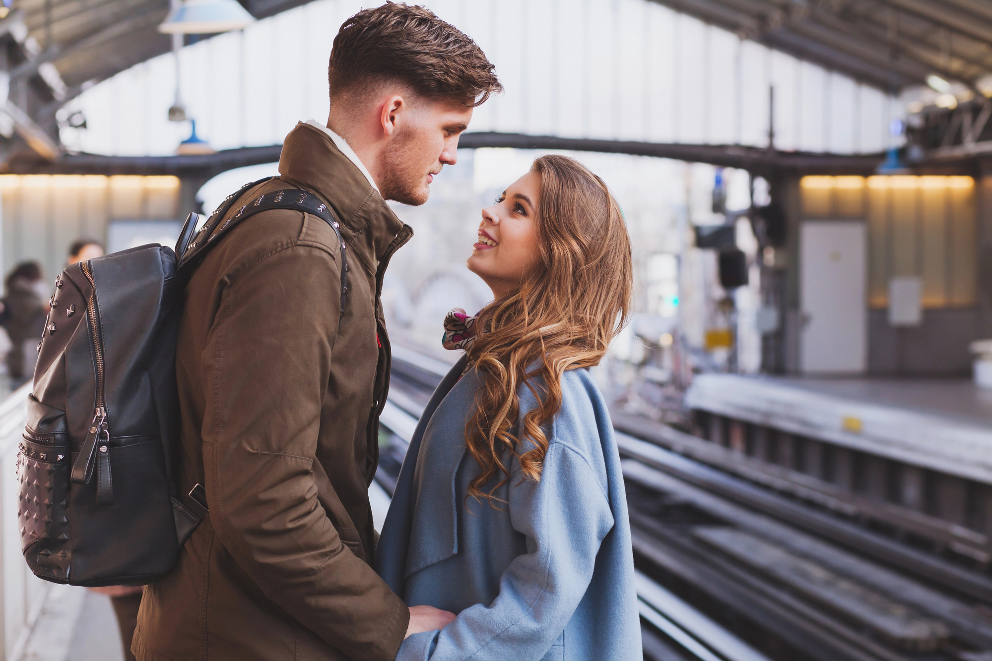 A man and woman stand close together at a train station, looking into each other's eyes. The man wears a brown jacket and a black backpack, while the woman is dressed in a light blue coat. They appear to be enjoying a tender moment amidst the busy setting.