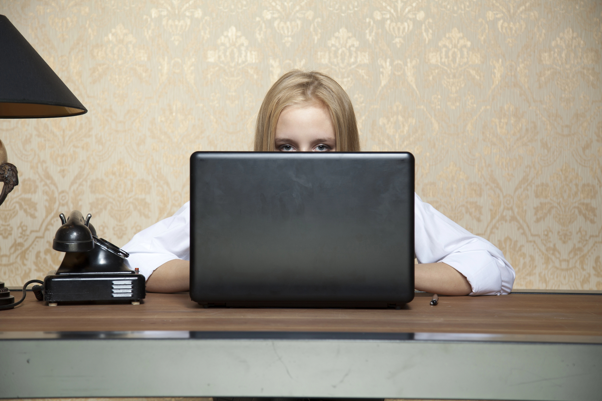 A person with blonde hair peeks over the top of a laptop, which is placed on a wooden desk. The background features a patterned wallpaper. An old-fashioned black telephone and a black desk lamp are also on the desk.