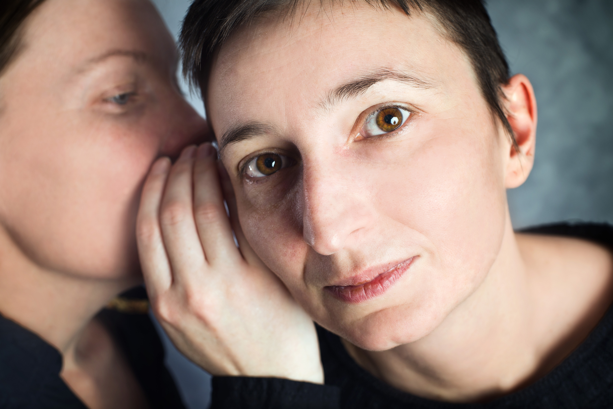 A close-up of two people sharing a secret. One person, who is out of focus, is whispering into the ear of the other, who has a serious expression and short hair, looking intently at the camera. The background is a soft, textured blue.