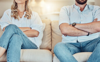 A man and a woman sit on a couch with their arms crossed, facing forward. Both are wearing light blue shirts and jeans. The background appears to be a softly lit living room, but their facial expressions are not visible as the image is cropped at their shoulders.