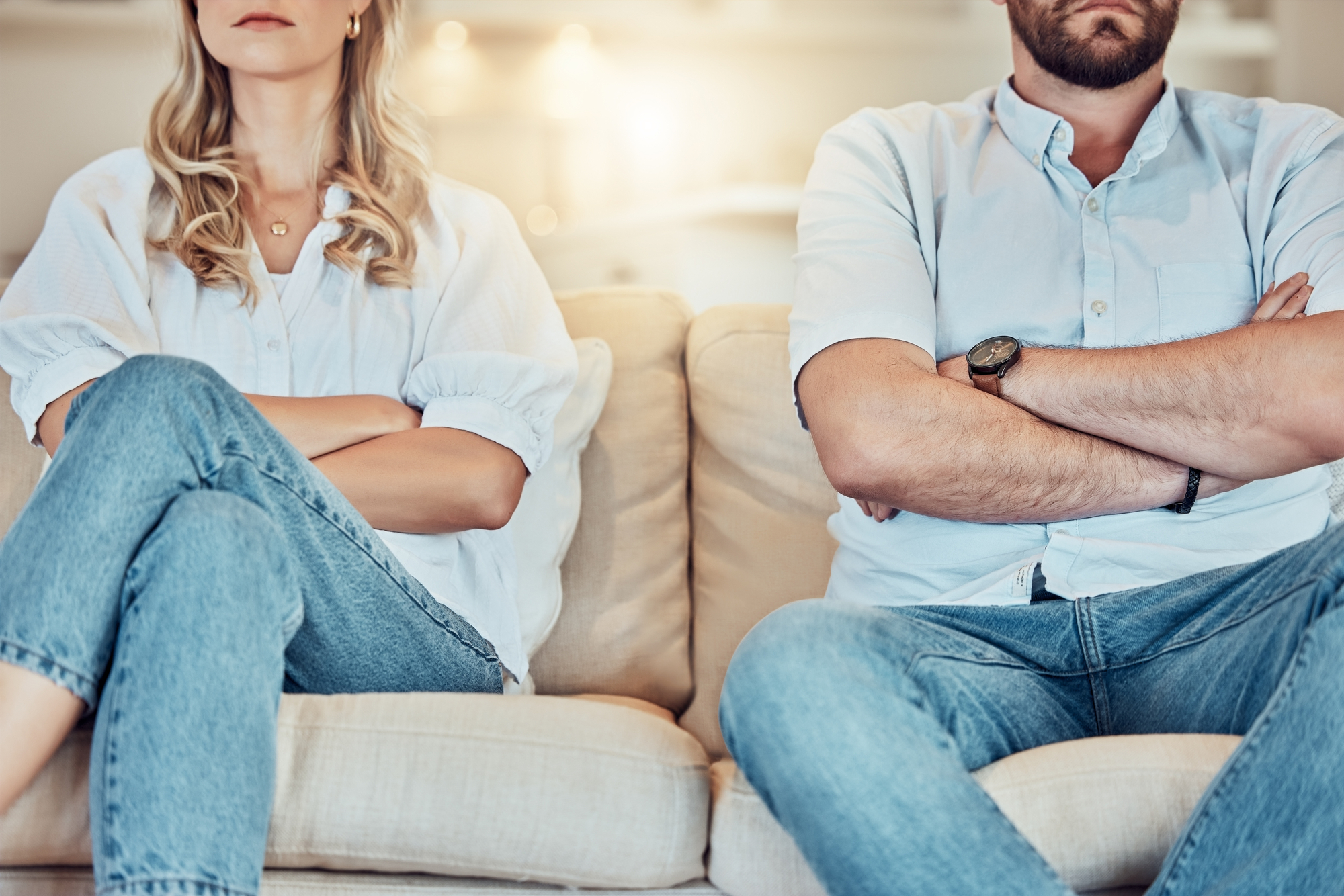 A man and a woman sit on a couch with their arms crossed, facing forward. Both are wearing light blue shirts and jeans. The background appears to be a softly lit living room, but their facial expressions are not visible as the image is cropped at their shoulders.