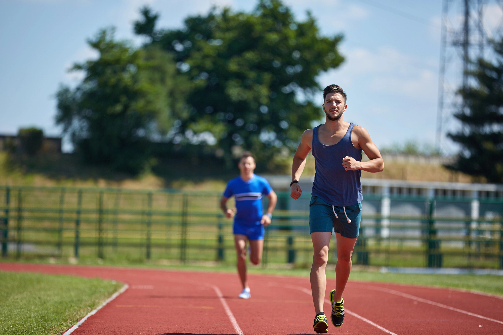 Two men are running on an outdoor track. The man in the foreground is wearing a grey tank top and blue shorts, while the man in the background is wearing a blue outfit. They are running on a red track with green grass and trees visible in the background.