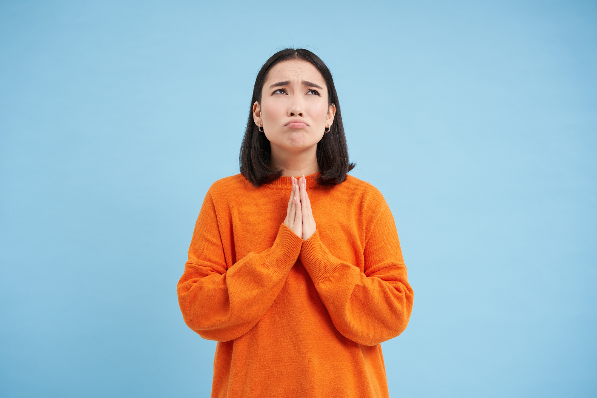 A person with shoulder-length dark hair is wearing an oversized orange sweater and holding their hands together in a pleading gesture with a sad facial expression. The background is a solid light blue.