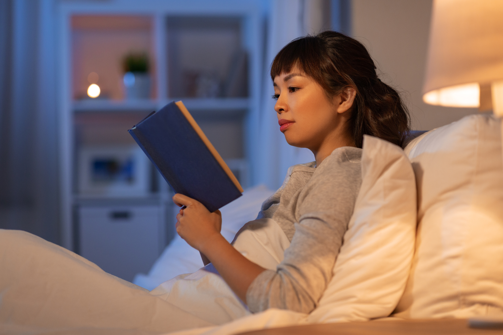 A woman sits in bed, comfortably propped up with white pillows, reading a book with a blue cover. She is wearing a gray long-sleeved top, and a lit lamp on a nightstand casts a warm, cozy glow. A shelf with decor and a candle is in the background.