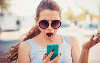 A woman with long hair tied back, wearing large round sunglasses and a light grey sleeveless top, is looking at her phone with an expression of shock or frustration. She is holding a turquoise phone in one hand and has her other hand raised in exasperation.