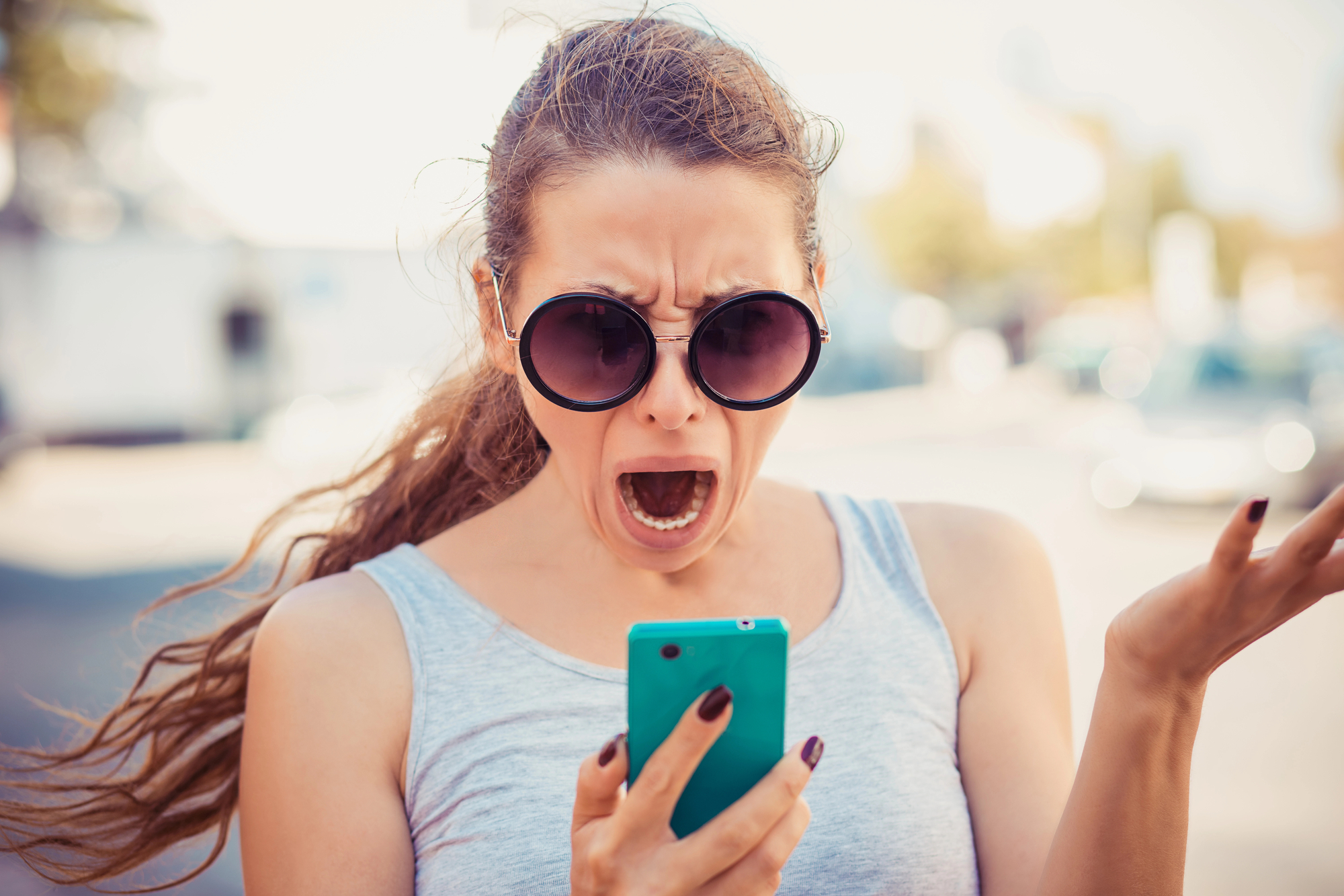 A woman with long hair tied back, wearing large round sunglasses and a light grey sleeveless top, is looking at her phone with an expression of shock or frustration. She is holding a turquoise phone in one hand and has her other hand raised in exasperation.