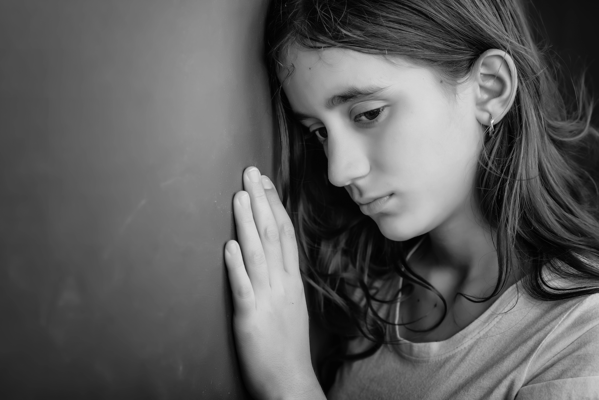 A black-and-white portrait of a young girl with long hair and a somber expression. She leans her forehead and hands against a wall, looking downwards. She wears a light-colored shirt and a small hoop earring in her left ear.