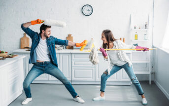 A man and a woman are playfully engaging in a mock sword fight in a bright, modern kitchen. The man wields a duster, and the woman uses a mop. Both are wearing casual clothes and cleaning gloves, and they appear to be having fun. The kitchen is clean and organized.