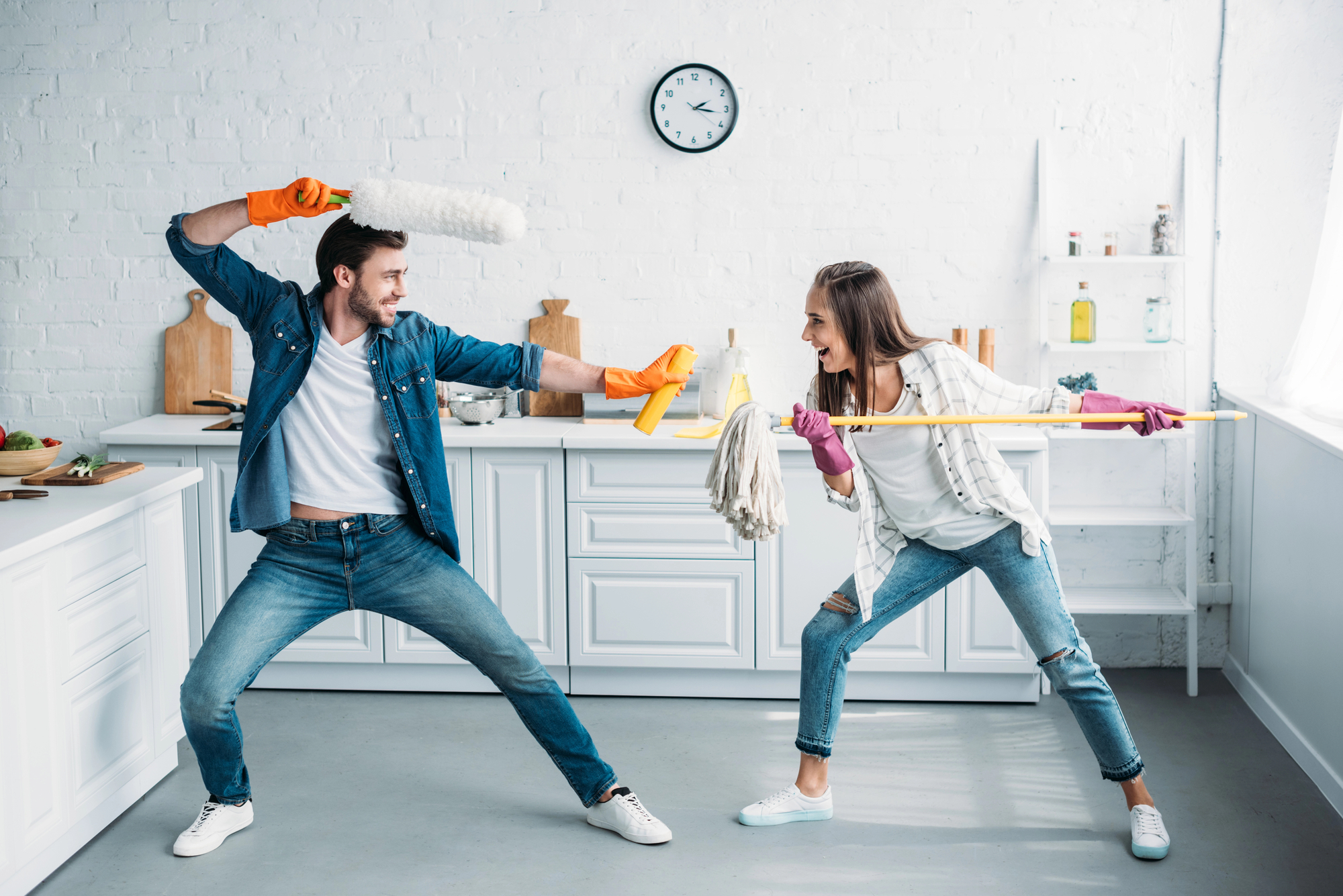 A man and a woman are playfully engaging in a mock sword fight in a bright, modern kitchen. The man wields a duster, and the woman uses a mop. Both are wearing casual clothes and cleaning gloves, and they appear to be having fun. The kitchen is clean and organized.