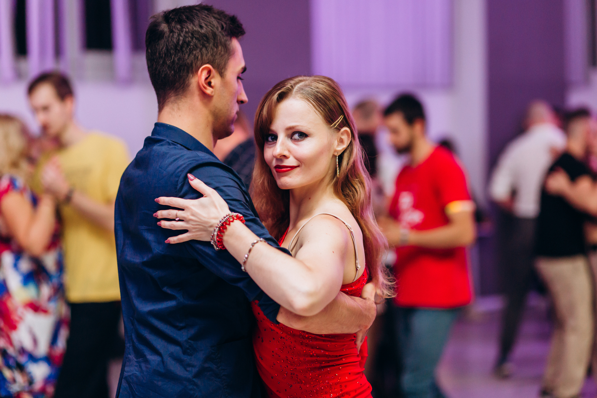 A man and woman are dancing closely in a brightly lit room, surrounded by other dancing couples. The woman, wearing a red dress with matched accessories, looks into the camera, while the man, in a dark shirt, faces away from it.