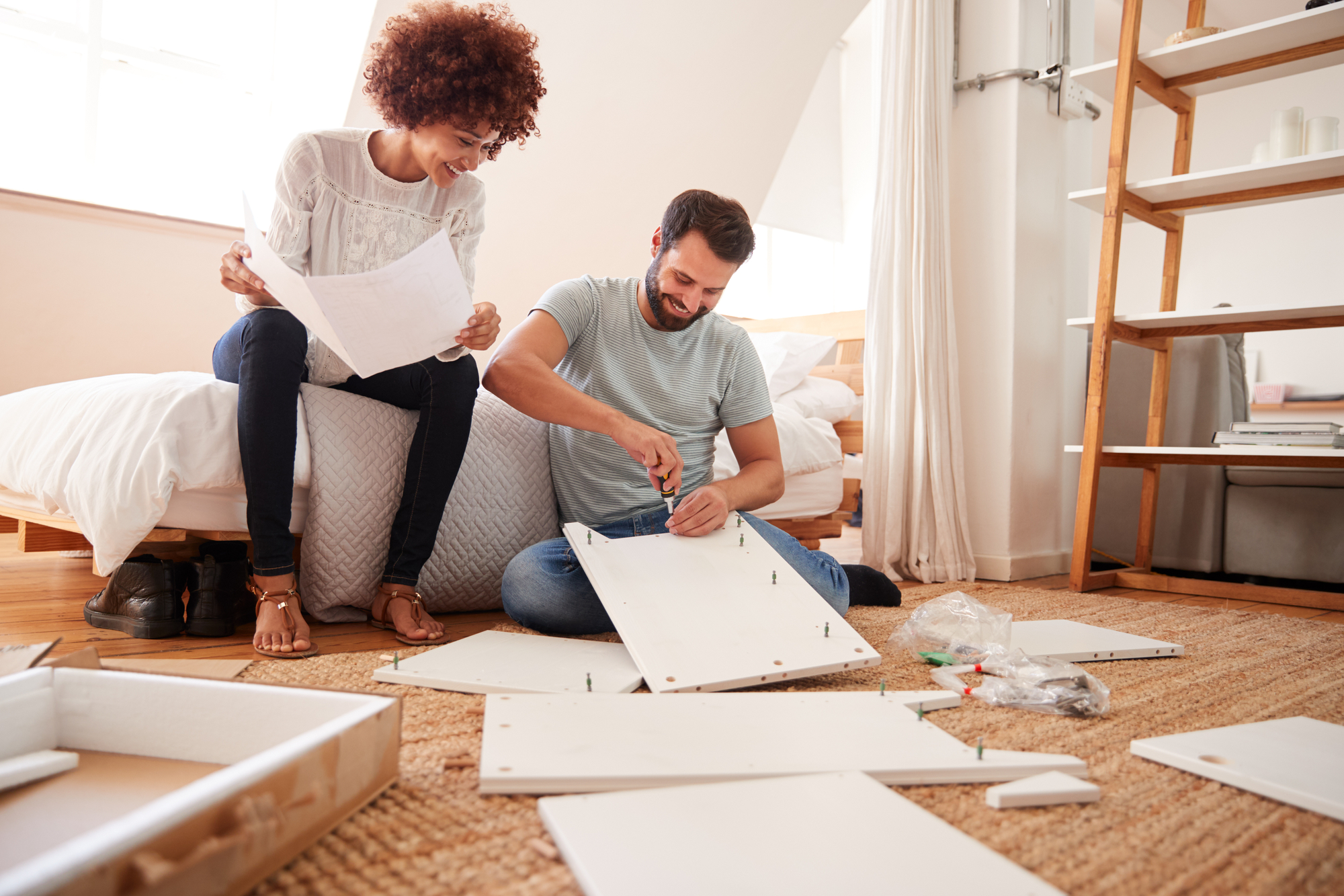 A woman and a man are assembling furniture in a bedroom. The woman is sitting on the bed, holding instruction papers and smiling, while the man is kneeling on the floor, using a screwdriver. Various furniture pieces and tools are scattered around them.