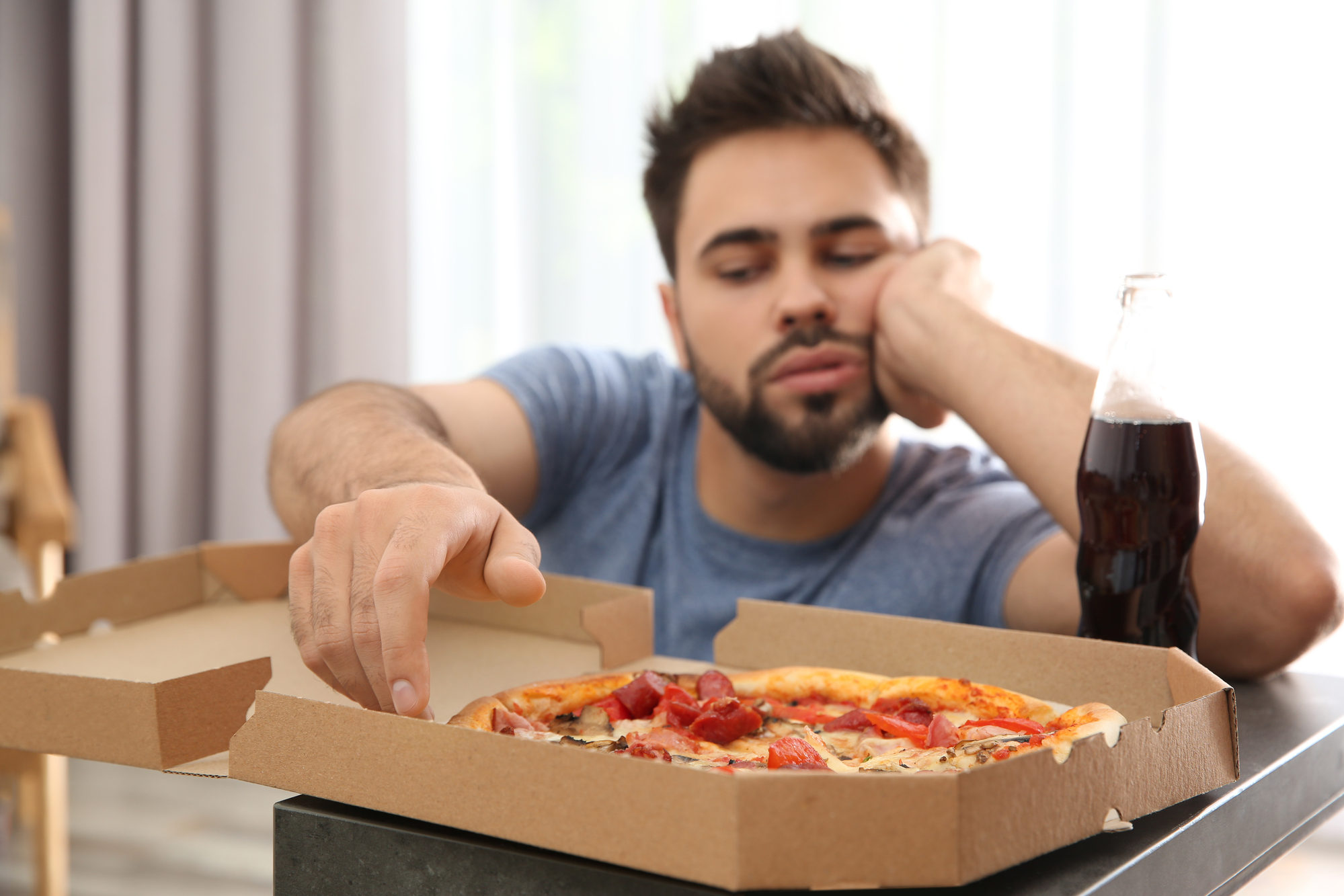 A man with a beard, wearing a blue shirt, sits at a table with an open pizza box and a bottle of soda. He looks uninterested as he rests his head on one hand and reaches for a slice of pizza with the other hand. The background is a bright room with curtains.