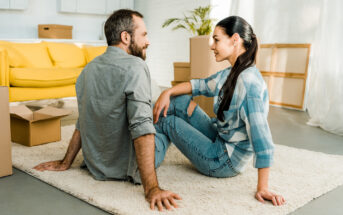 A man and a woman, both casually dressed, are sitting on a white carpet in a brightly lit room. The room has several moving boxes and a yellow couch in the background. Both individuals are smiling at each other, suggesting they are in the process of moving in together.